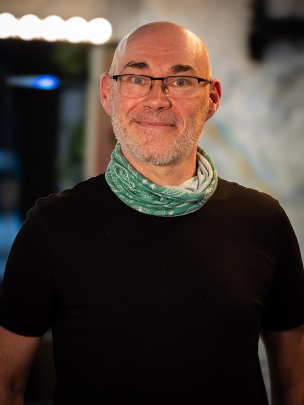 A middle-aged man with glasses, a bald head, and a short grey beard wears a black shirt and a green patterned neck gaiter, standing indoors in soft lighting.