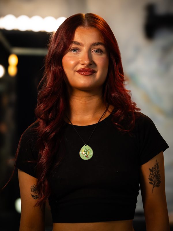 A woman with long red hair, wearing a black crop top and a round pendant necklace, stands indoors with soft lighting and blurred background.