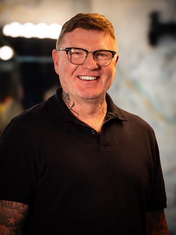 A man with short hair, glasses, and visible tattoos is smiling, wearing a black polo shirt, standing indoors with blurred lights and a wall in the background.