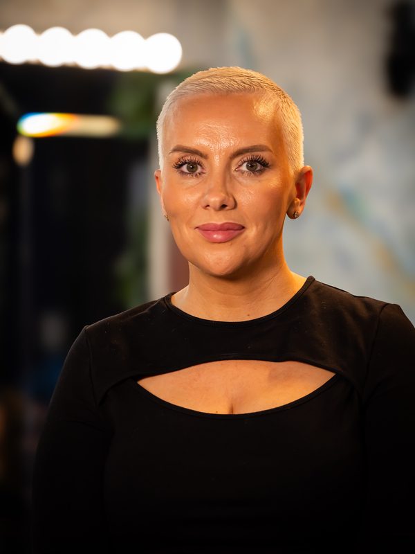 A woman with short platinum blonde hair and a black top poses indoors, looking directly at the camera with a neutral expression.