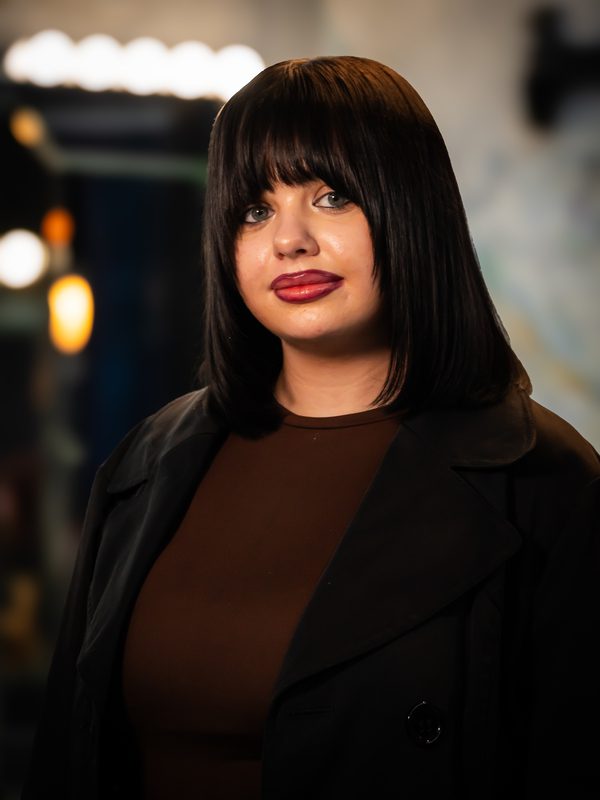 A woman with straight black hair and bangs, wearing a brown top and black coat, stands indoors with soft, blurred lights in the background.