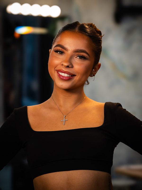 A young woman with styled hair and makeup smiles at the camera, wearing a black top and a cross necklace, standing indoors with blurred lights in the background.