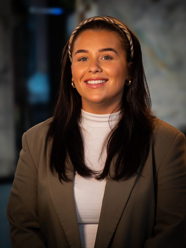 A woman with long dark hair wearing a headband, white turtleneck, and taupe blazer smiles at the camera in an indoor setting.