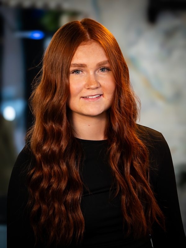 A young woman with long, wavy red hair and fair skin wearing a black top, smiling in front of a blurred indoor background.