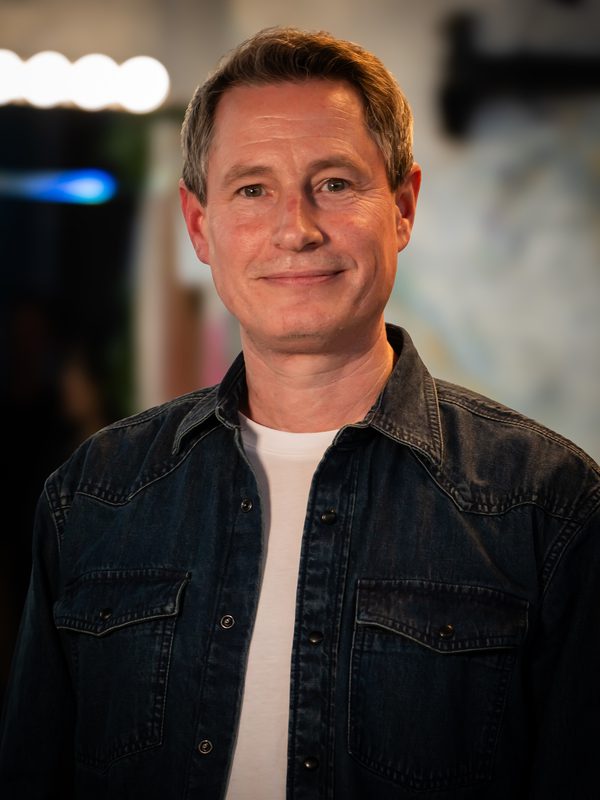 A middle-aged man with short brown hair, wearing a dark denim shirt over a white t-shirt, stands indoors and smiles at the camera.