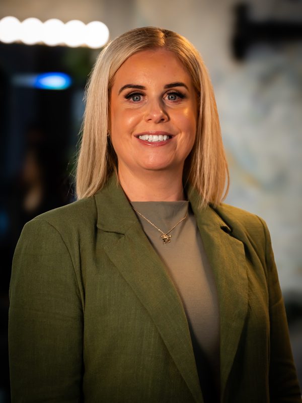 A woman with straight blonde hair wearing a green blazer and gray top stands indoors, smiling at the camera.