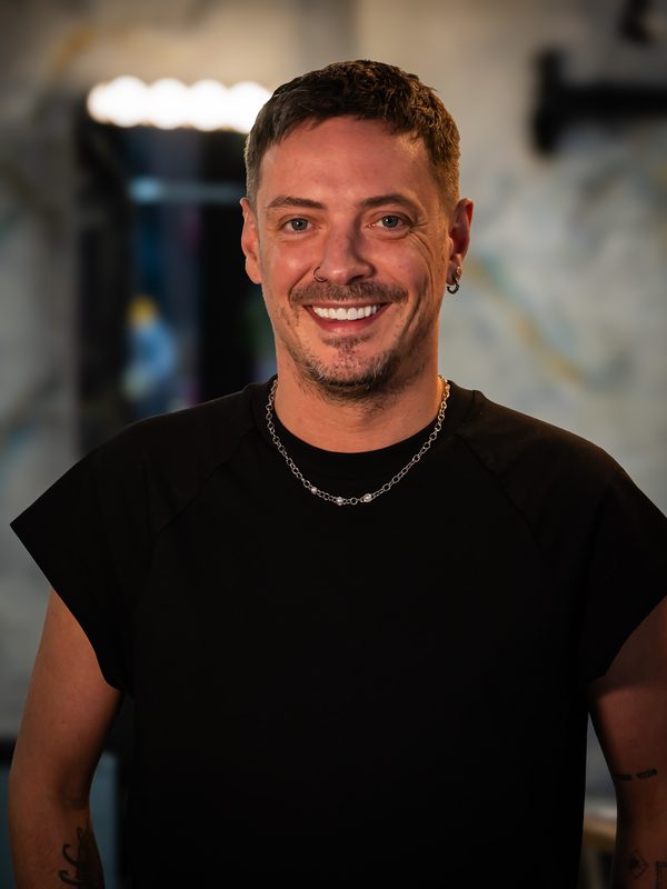 A person with short brown hair, facial hair, and earrings is smiling while wearing a black shirt and a chain necklace, standing indoors.