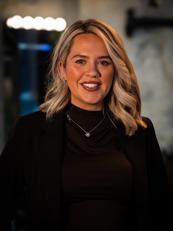A woman with shoulder-length blonde hair, wearing a black top and blazer, smiles at the camera in a dimly lit indoor setting.