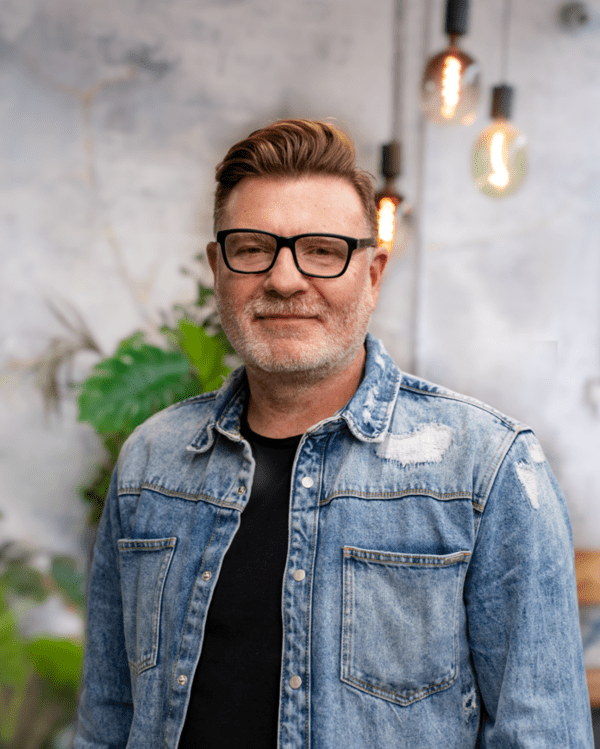 A man with glasses and a beard wears a denim jacket over a black shirt, standing indoors with plants and hanging lights in the background.